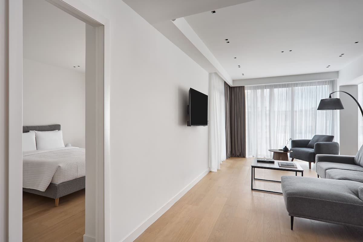 A modern hotel room with a bed, a sofa, a coffee table, a TV mounted on the wall, and a large window with curtains.