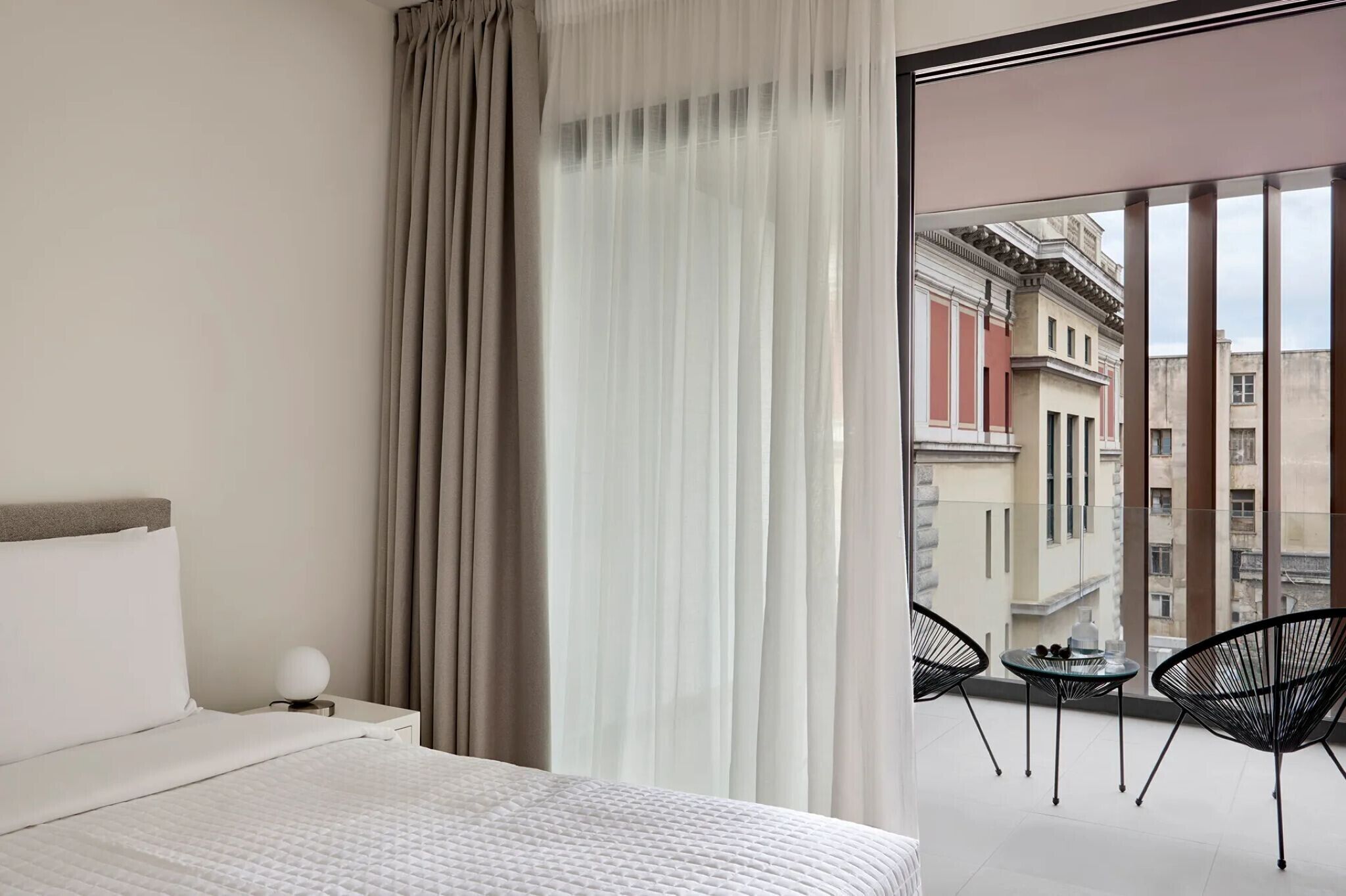 A hotel room with a bed, a nightstand with a lamp, a balcony with chairs, and a view of buildings.