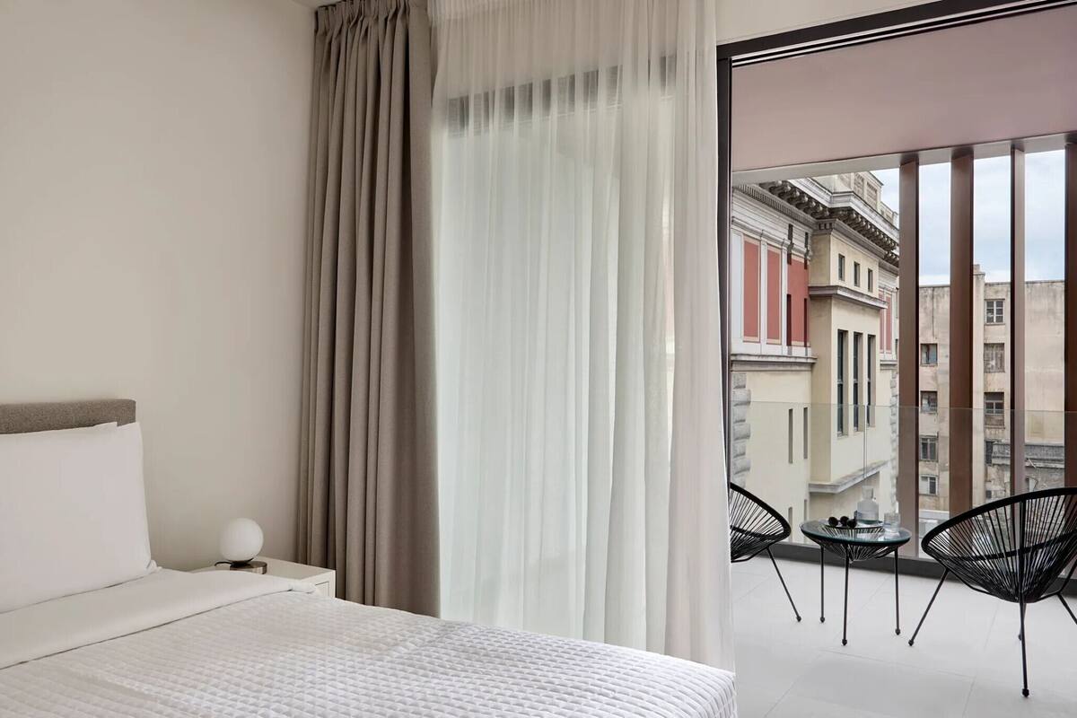 A hotel room with a bed, a nightstand with a lamp, a balcony with chairs, and a view of buildings.