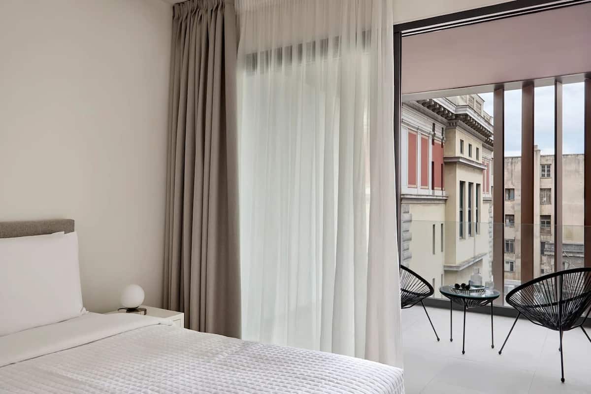 A hotel room with a bed, a nightstand with a lamp, a balcony with chairs, and a view of buildings.