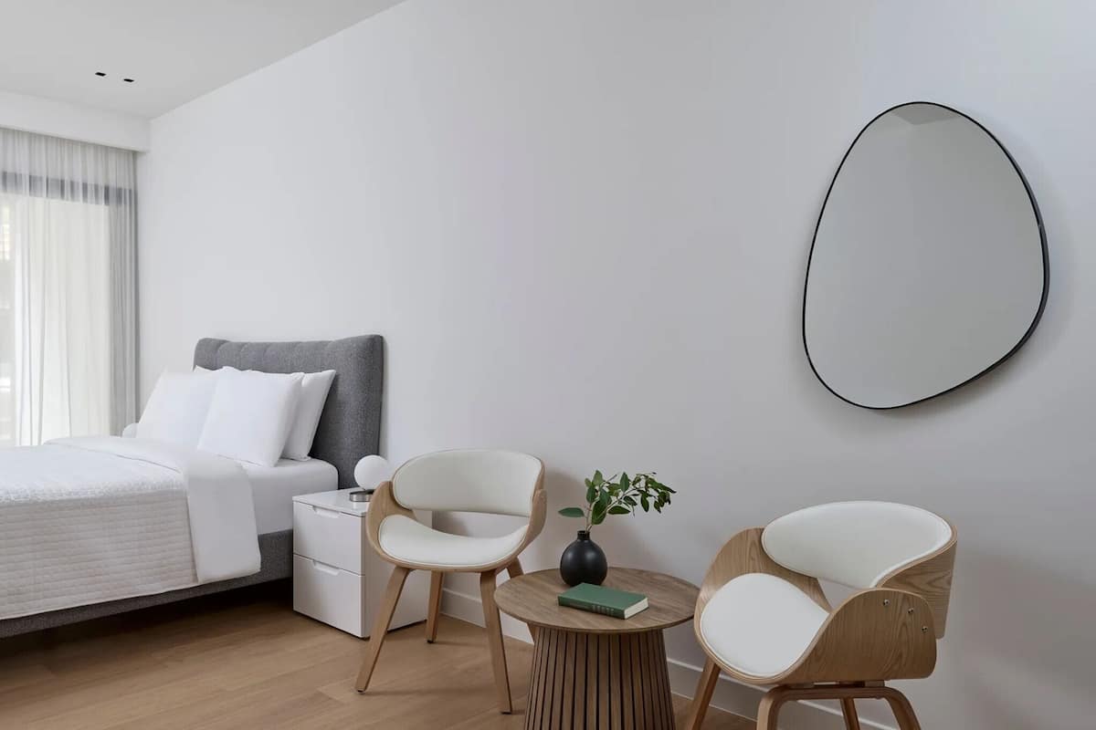 A modern hotel room with a bed, bedside table, two chairs, a small round table with a plant, and a large mirror on the wall.