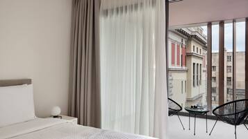 A hotel room with a bed, a nightstand, a round table with chairs, and a view of buildings through large windows.