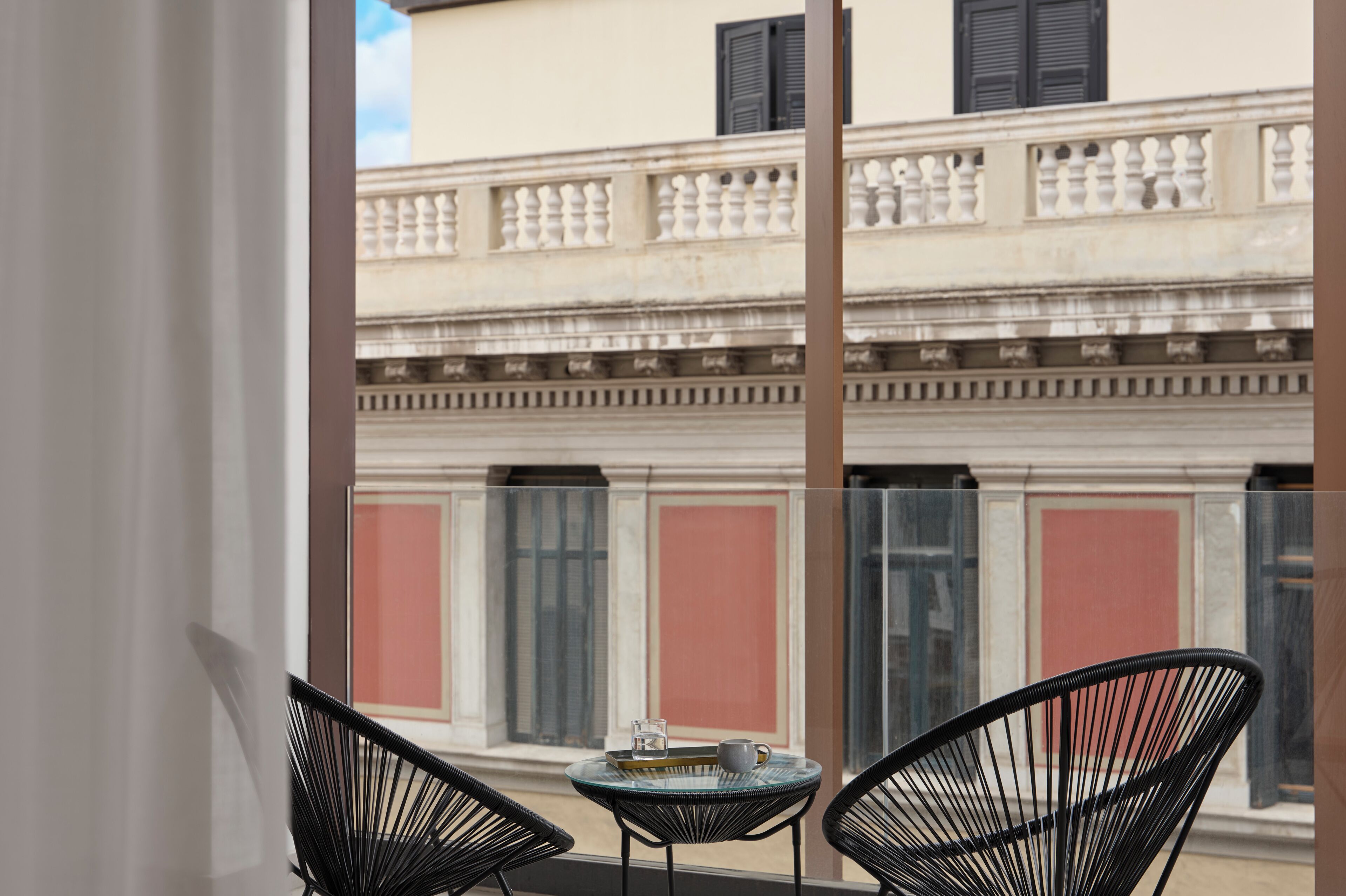 A balcony with two chairs and a small table, overlooking a building with classical architecture and red shutters.