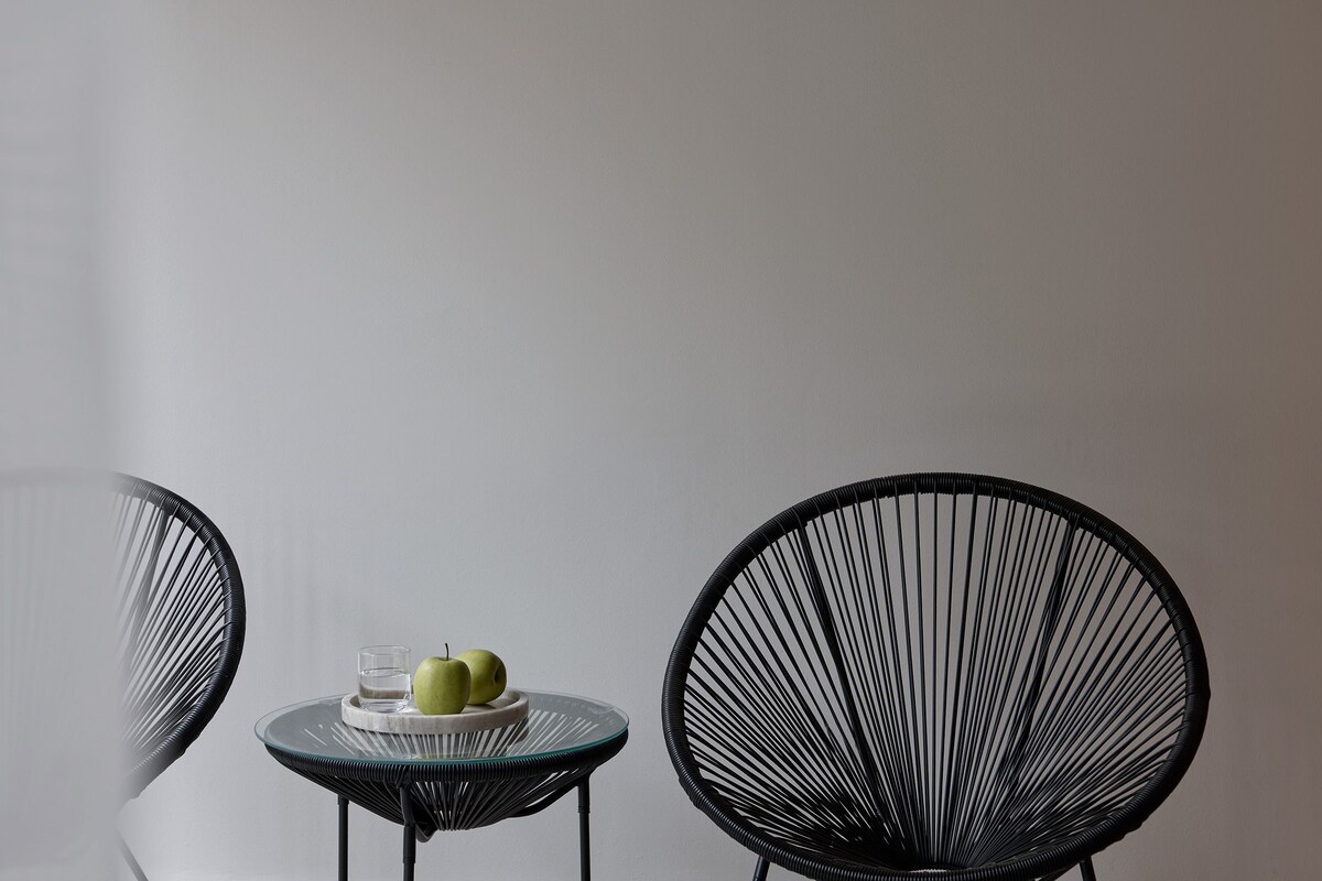 Two black wireframe chairs, a small round table with a glass and two apples, and a plain wall.