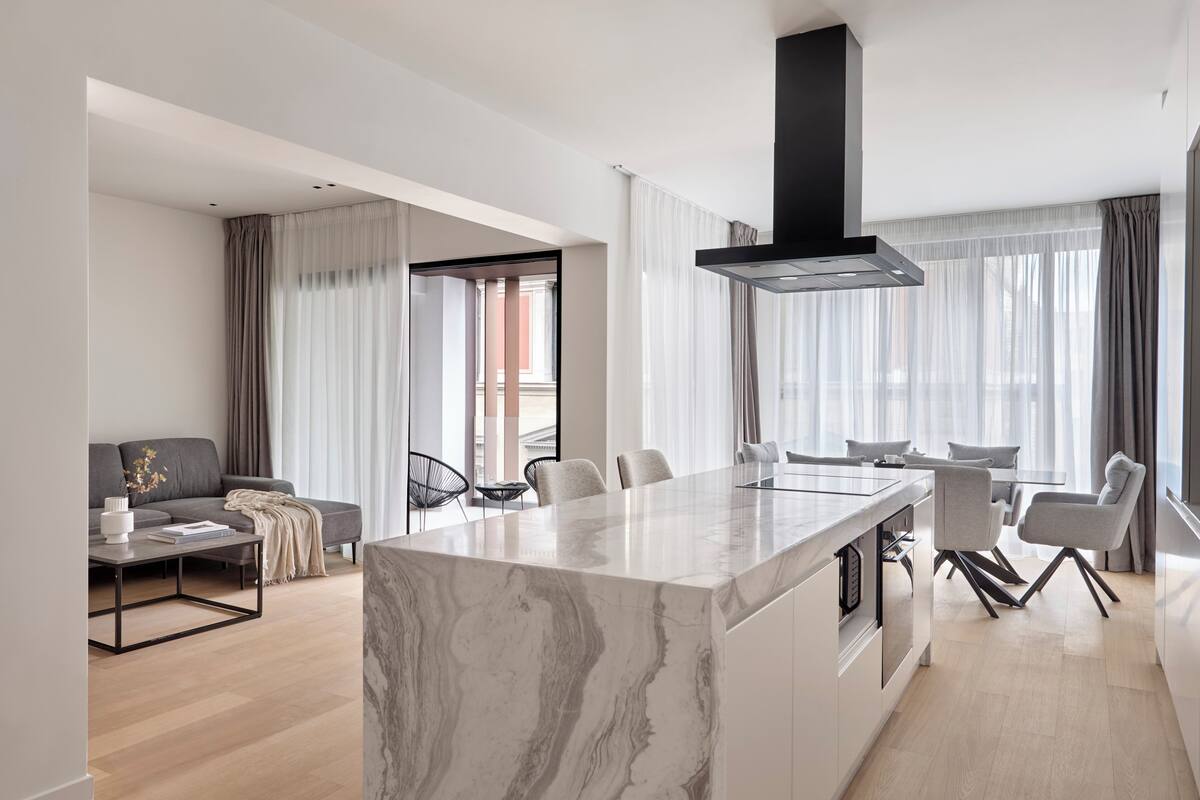 A modern living space with a marble island, a black range hood, and a dining area with chairs.