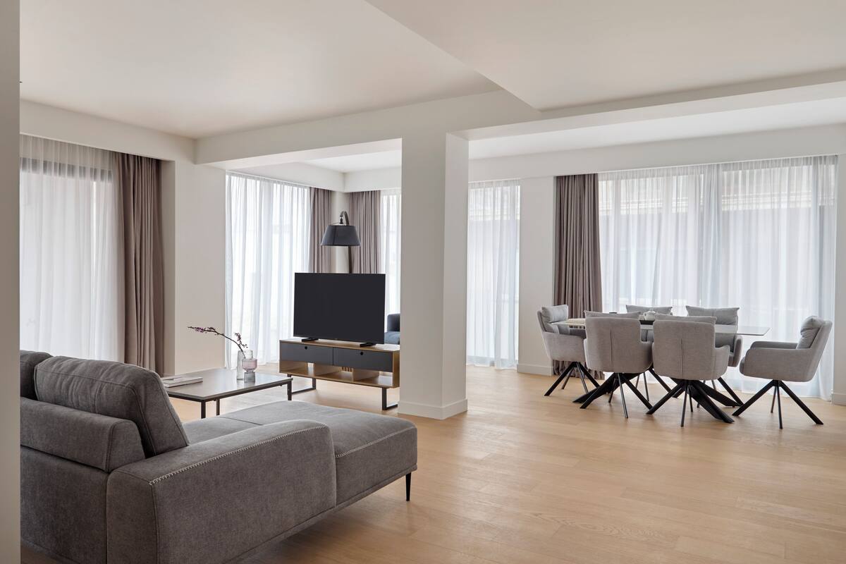 A modern living room with a grey sofa, a TV, a dining area with a table and chairs, and large windows with curtains.