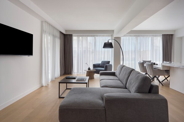 Un salon moderne avec un canapé d’angle gris, un téléviseur à écran plat fixé au mur, une petite table ronde et un coin repas avec une table et des chaises blanches.