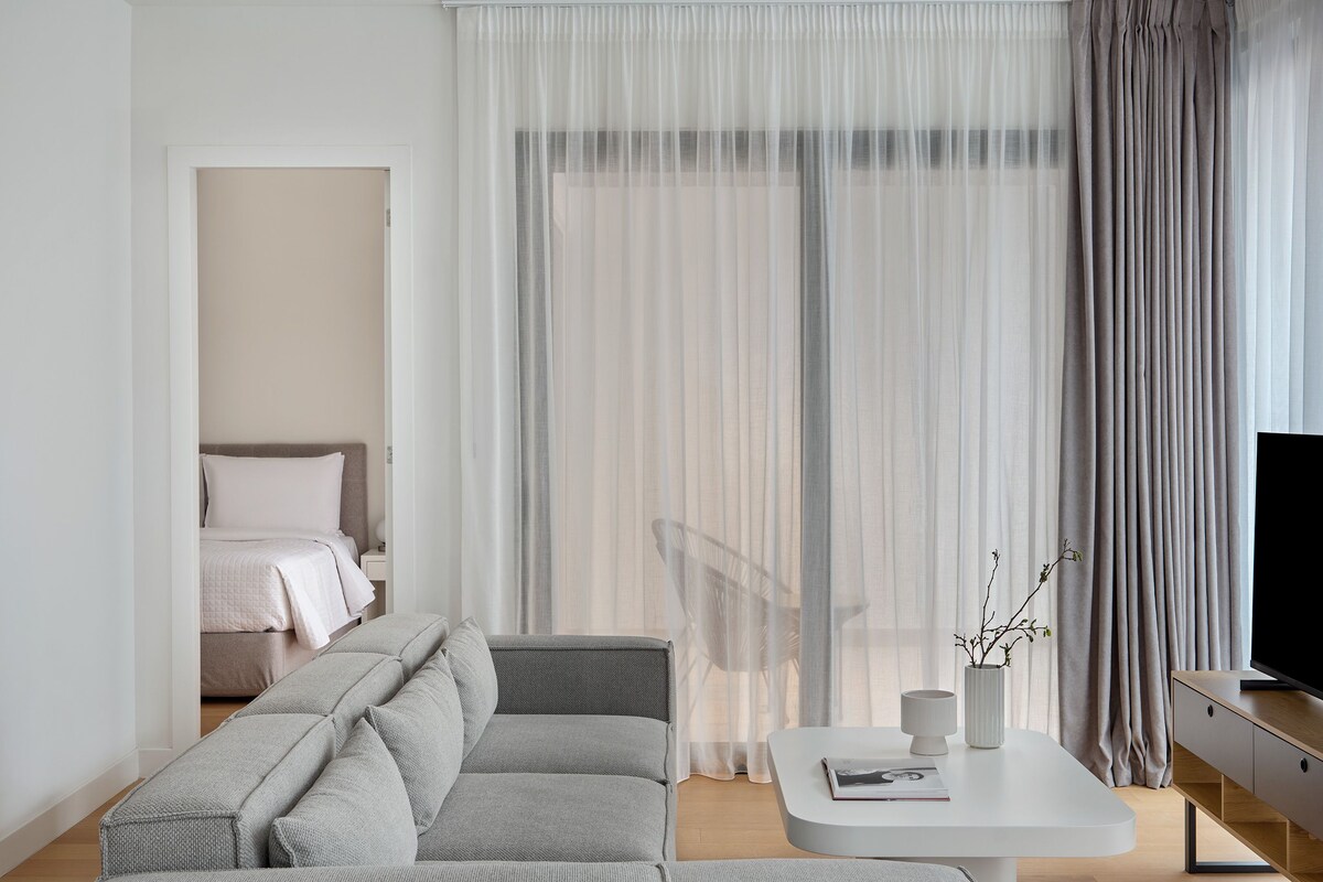 A modern hotel room with a grey sofa, a white coffee table, a TV on a wooden stand, and sheer curtains.