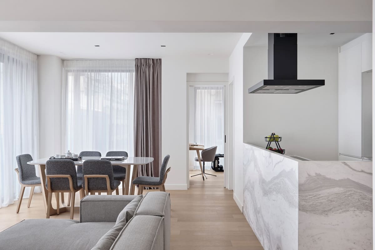 A modern living room with a dining area, a kitchen with a black range hood, and a marble countertop.