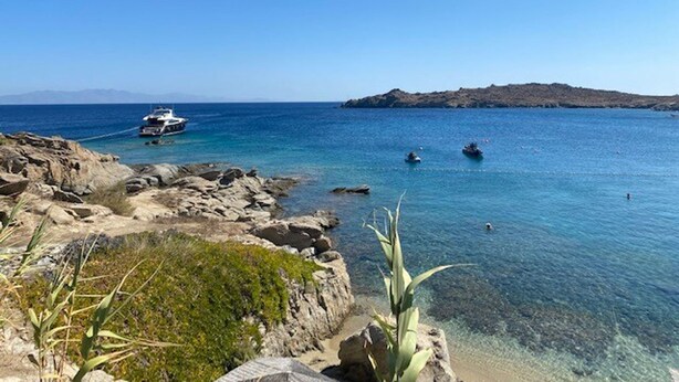 A rocky shoreline with clear blue water, a boat anchored near the shore, and a distant island. - Smart  4 pers. apar. in Costa Ilios resort Myconos (Mikonos)