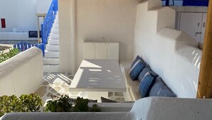 A covered outdoor seating area with a table and cushions, a staircase, and a view of a white building with blue accents. - Smart  4 pers. apar. in Costa Ilios resort Myconos (Mikonos)