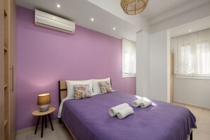 A bedroom with a purple bedspread, two pillows, a bedside lamp, and two rolled towels on the bed. - Levanta Cozy One-Bedroom Apartment in Kalavarda (Kalavarda)