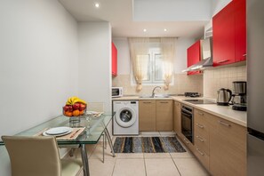 A compact kitchen with a glass dining table, a washing machine, and red cabinets. - Levanta Cozy One-Bedroom Apartment in Kalavarda (Kalavarda)