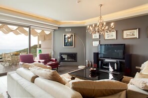 Ein modernes Wohnzimmer mit einer großen Couch, roten Sesseln, einem Kamin, einem Fernseher und Blick ins Freie.