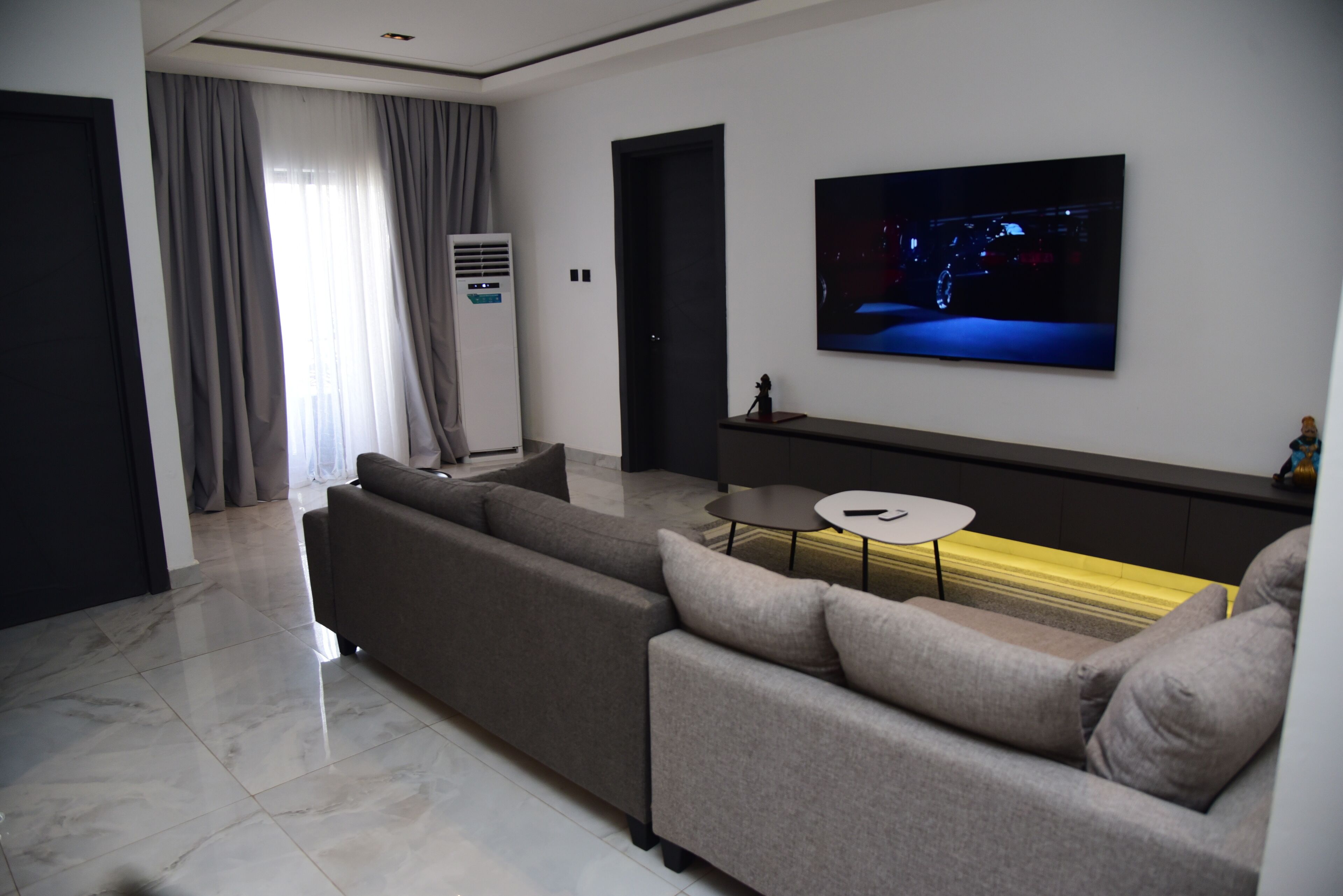 A modern living room with a large flat-screen TV mounted on the wall, a grey sofa, a small round coffee table, and a large window with curtains.