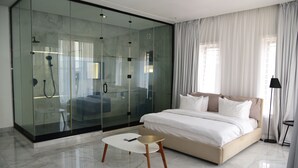 A modern bedroom with a glass-enclosed walk-in closet, a large bed, and a small white table with a phone. - Hotel De Alommah Kubwa Abuja Nigeria (Abuja)