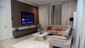 A modern living room with a large flat-screen TV mounted on the wall, a beige sofa with orange cushions, a white coffee table, and a minimalist design. - Hotel De Alommah Kubwa Abuja Nigeria (Abuja)