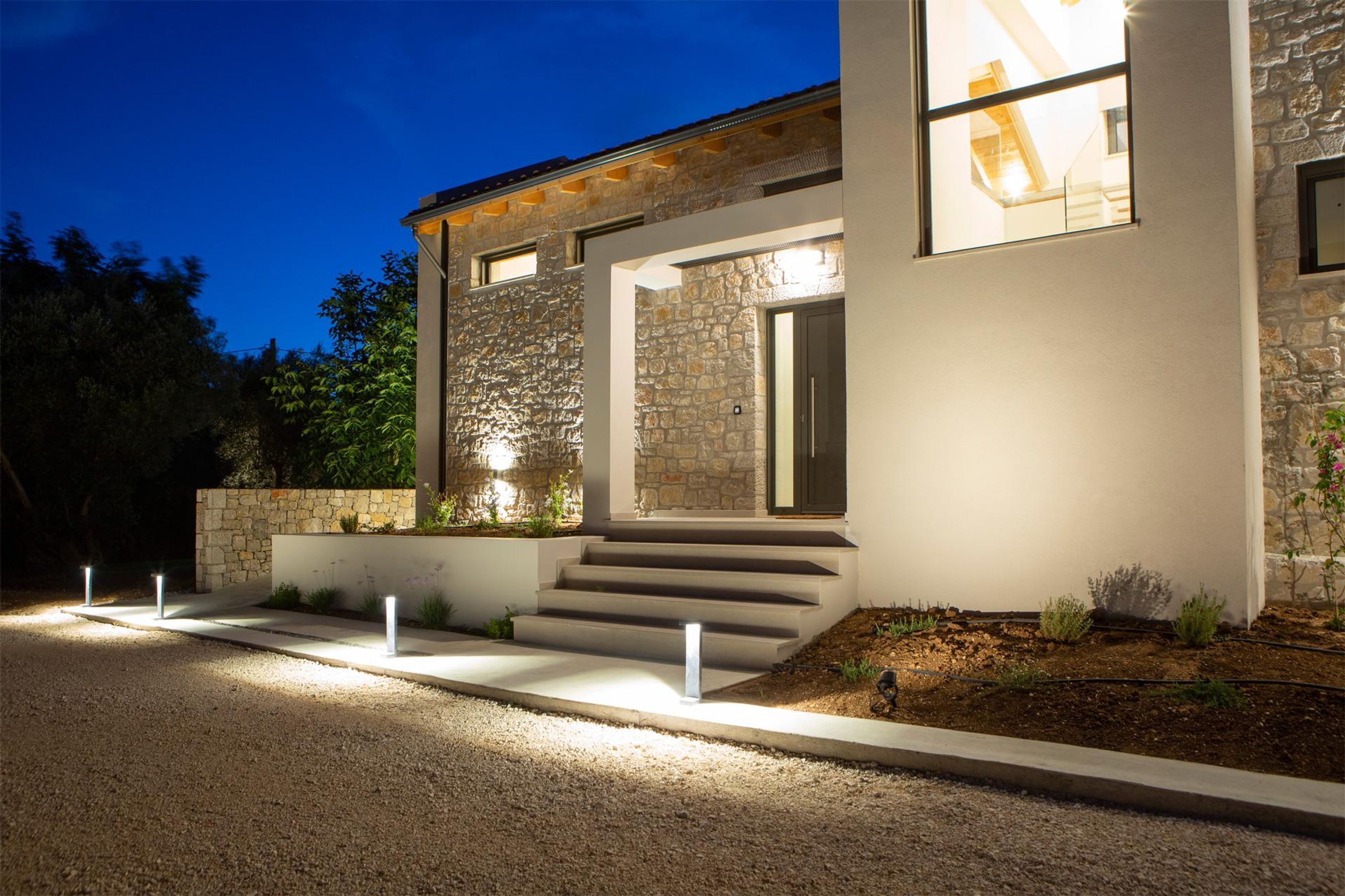 A modern house with a stone facade, a well-lit entrance, and a gravel driveway.