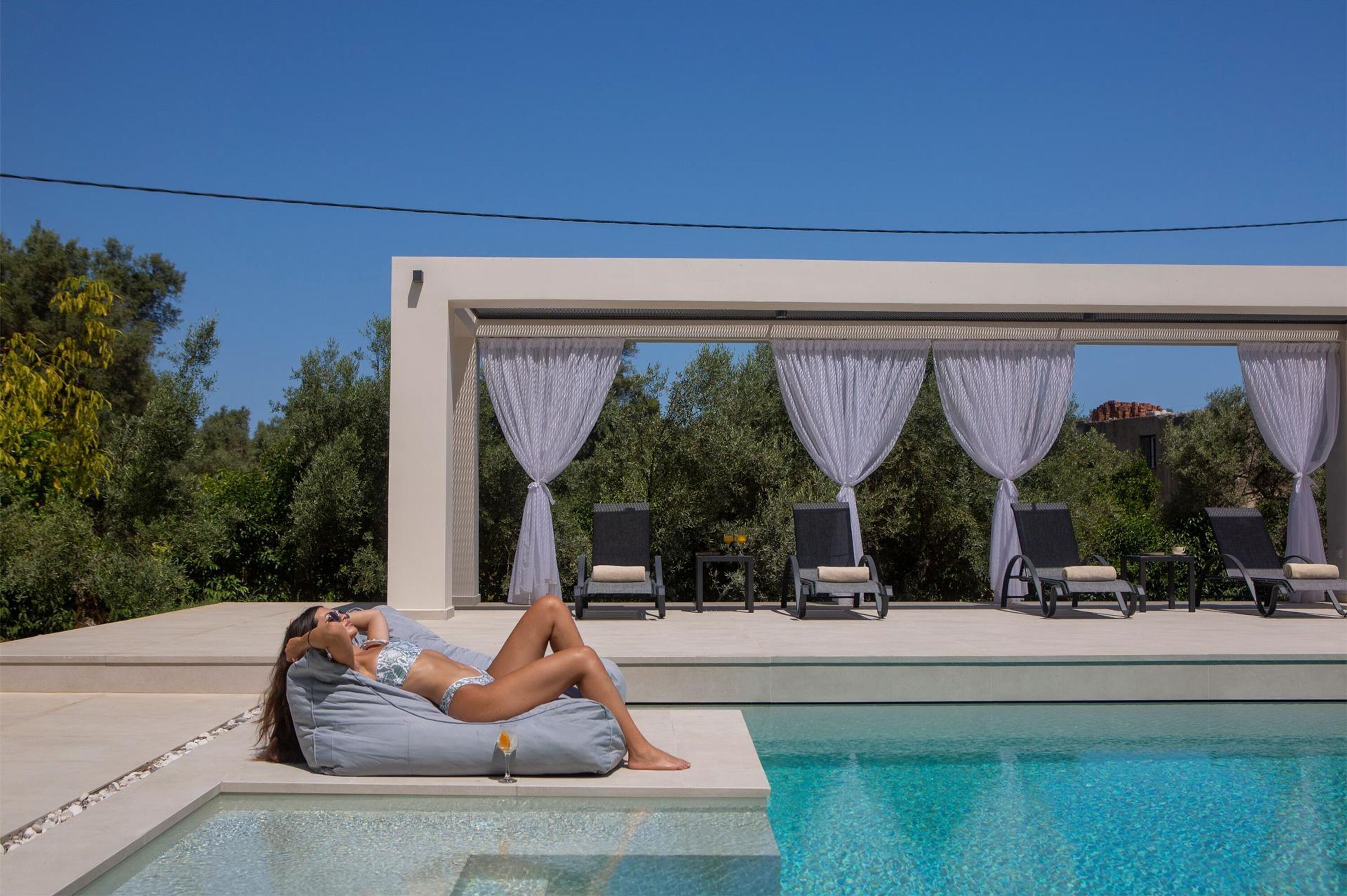 A poolside area with a person relaxing on a cushion, a covered seating area with chairs, and a clear blue pool.