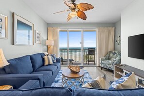 A living room with a blue sofa, a coffee table, a ceiling fan, and a view of the ocean through large windows. - Celadon 202 | Gulf-Front Views, 2 Pools, Sleeps 8 (Panama City Beach)