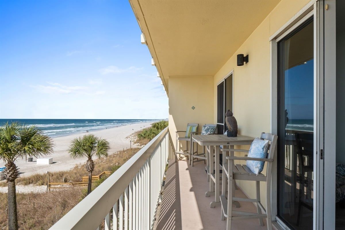 A balcony with a view of the beach, a table with chairs, and a door leading to the interior.