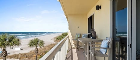 A balcony with a view of the beach, a table with chairs, and a door leading to the interior.