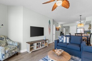 A living room with a TV, sofa, and a ceiling fan. - Celadon 202 | Gulf-Front Views, 2 Pools, Sleeps 8 (Panama City Beach)