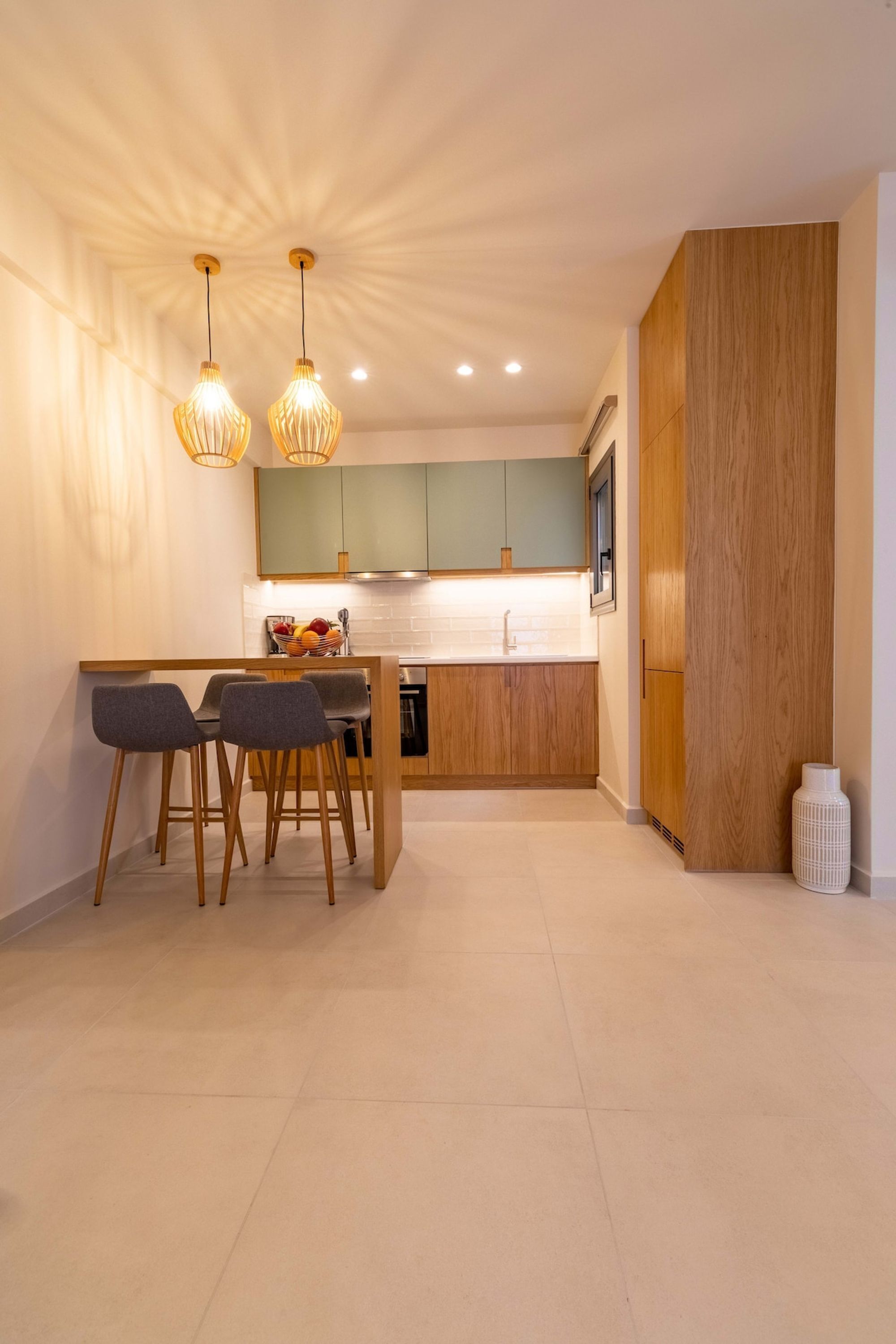 Eine moderne Küche mit Holzausstattung, einer zentralen Kochinsel mit Sitzgelegenheiten, Pendelleuchten und einem weißen Blumenvasen auf dem Boden.