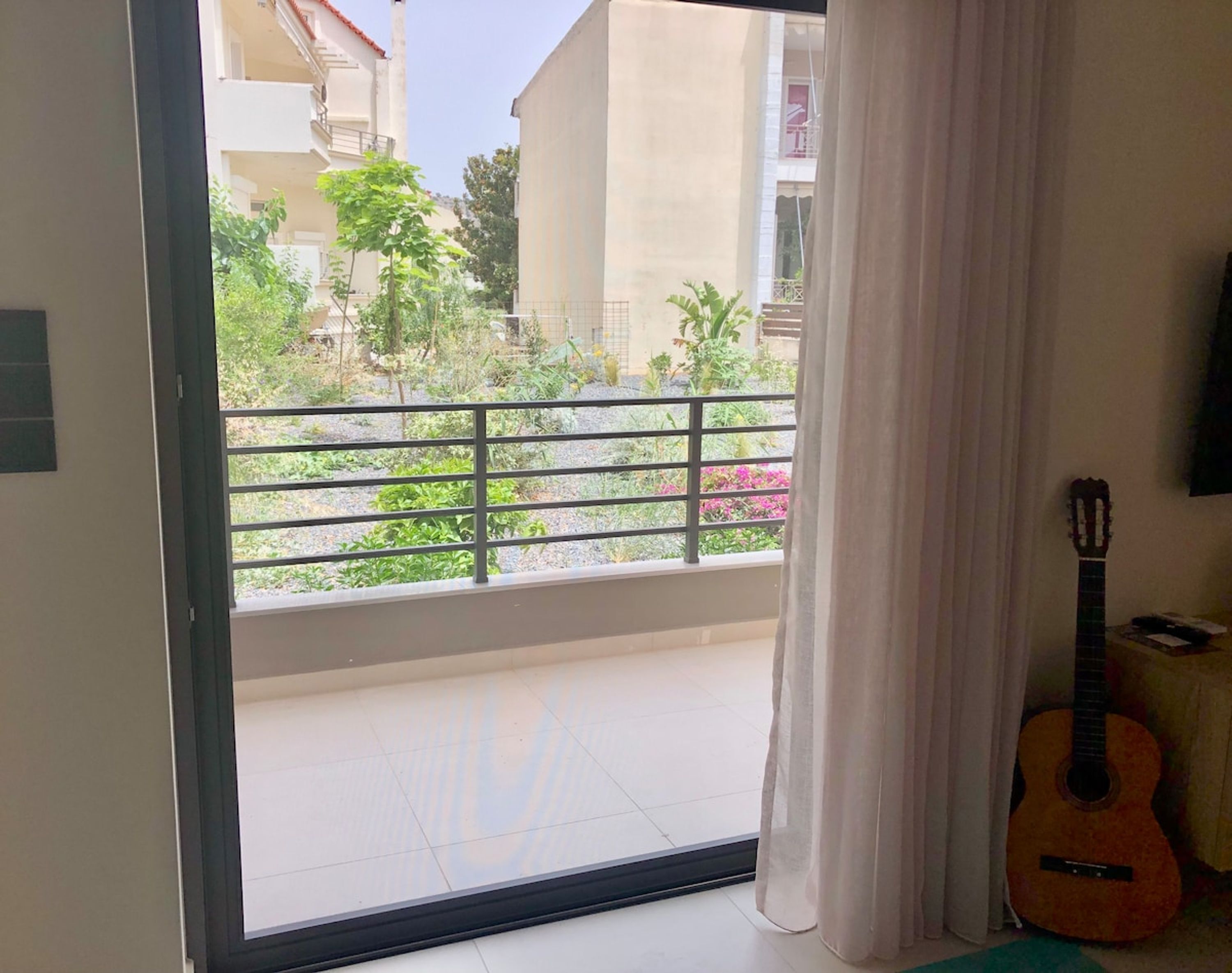 A balcony with a view of a garden and buildings, a guitar leaning against the wall, and a curtain partially drawn.