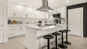 Een moderne keuken met witte kasten, een centraal eiland met barkrukken, een roestvrijstalen afzuigkap en een magnetron boven het eiland.