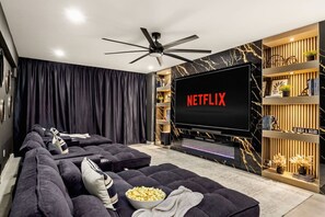 Ein modernes Wohnzimmer mit einem großen Flachbildfernseher, der 'NETFLIX' zeigt, einer dunkelgrauen Sofagarnitur, einem Deckenventilator und einem dekorativen Regal mit Pflanzen und Büchern.