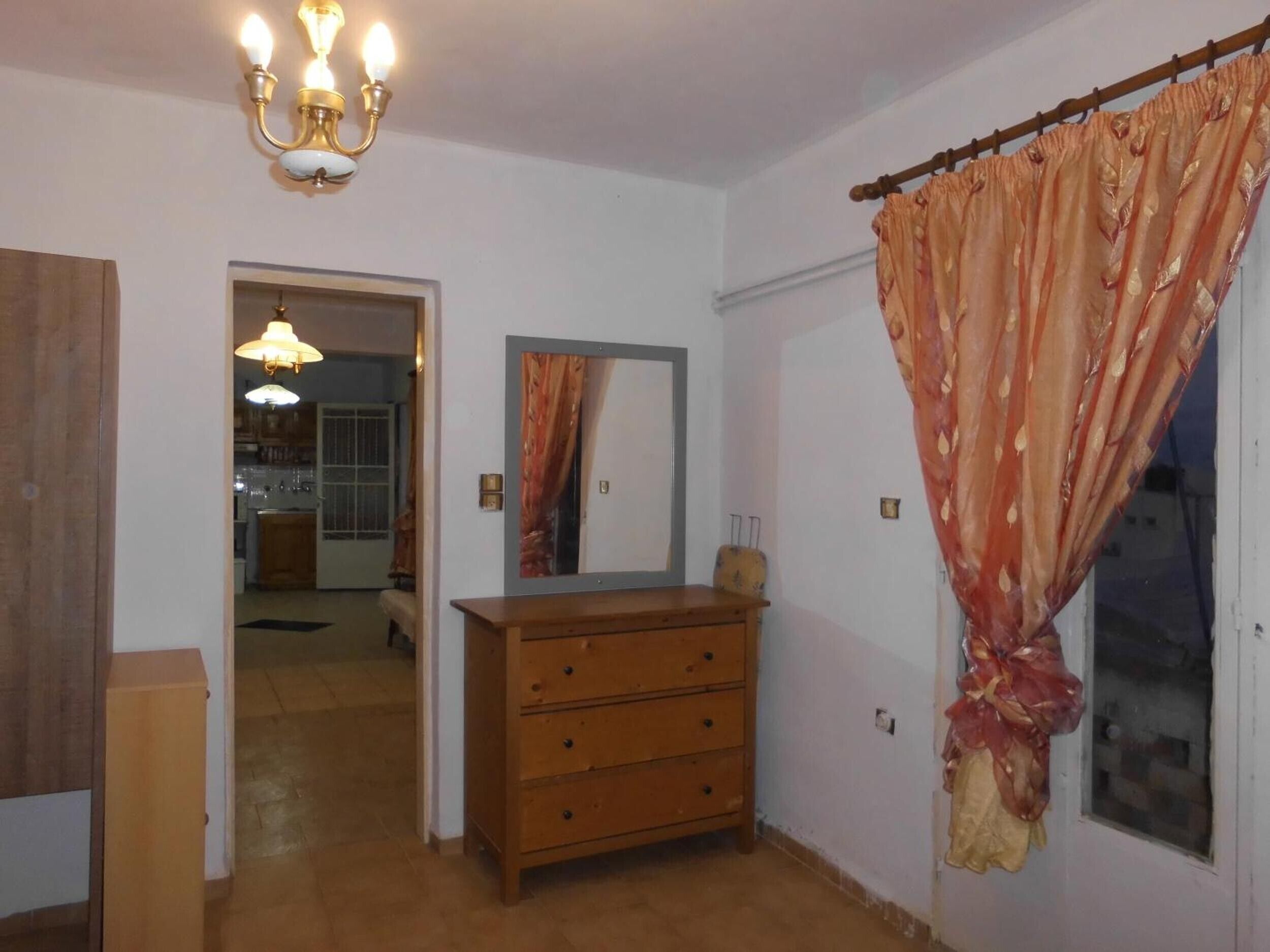 A room with a wooden dresser, a mirror, a chandelier, and a door leading to a kitchen.