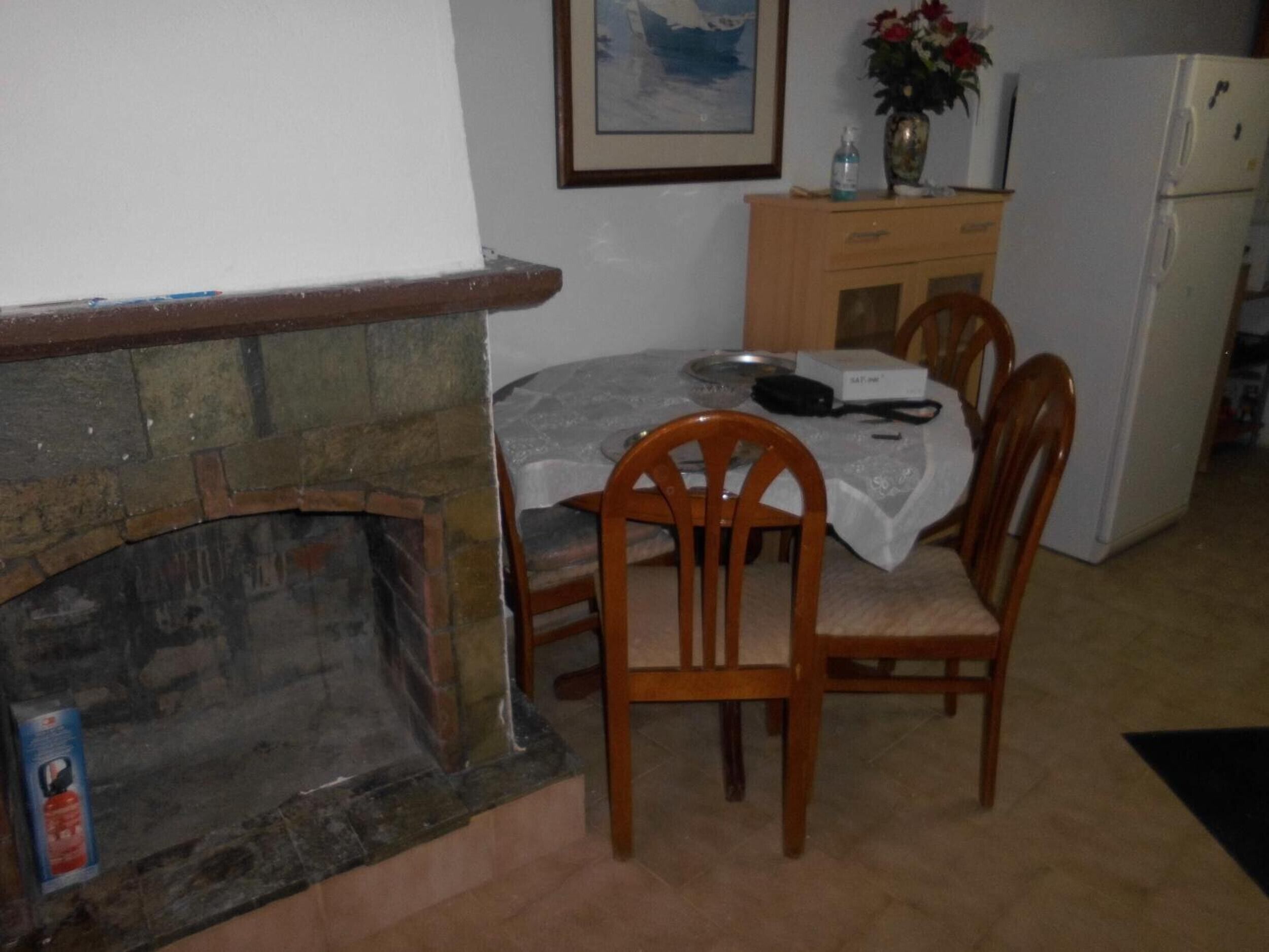 Ein Essbereich mit Kamin aus Stein, Holzstühlen, einem Tisch mit Tischdecke, einem Kühlschrank und einer Blumenvase auf einem Schrank.