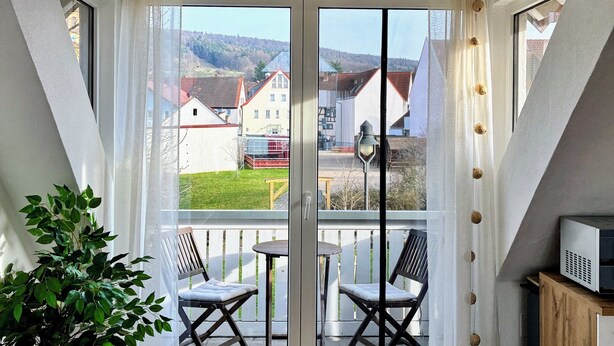 Ein Balkon mit Glastür, einem Blumentopf, zwei Stühlen und einem kleinen Tisch.