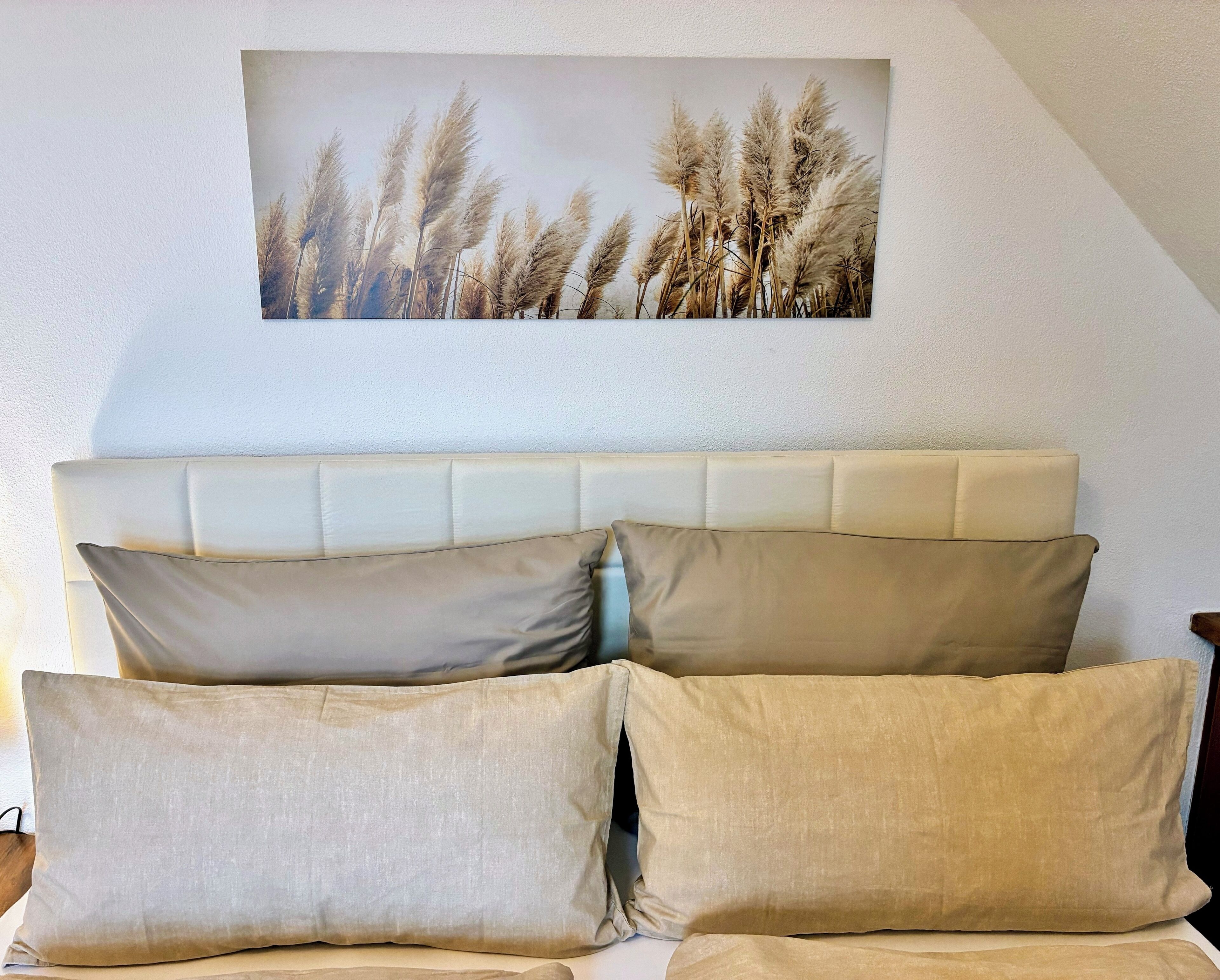 Une chambre avec une tête de lit blanche, des coussins de couleur beige et un tableau représentant des plumes d’herbe des pampas, accroché au mur.