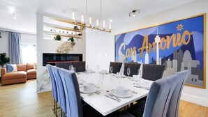 Een moderne eetruimte met een witte tafel voor zes personen, blauwe bekleden stoelen en een grote muurschildering van 'San Antonio' op de wand.