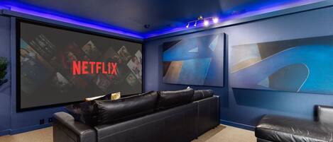 A home theater room with a large screen displaying 'NETFLIX', a black leather couch, and abstract wall art.