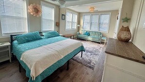 Ein Schlafzimmer mit Bett, Deckenventilator, Fenster mit Jalousien, einer Lampe und einem kleinen Tisch.