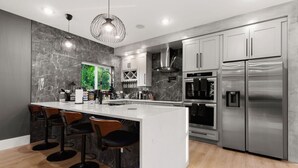 A modern kitchen with a marble backsplash, stainless steel appliances, and a central island with bar stools. - City Views Rooftop Karaoke, Pool, Hot Tub, Games (San Antonio)