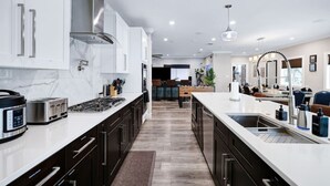 A modern kitchen with dark cabinetry, a central island with a sink, and a dining area in the background. - Stylish 6BR/5BA, DT, Hot Tub, Games, Sleeps 20+ (San Antonio)
