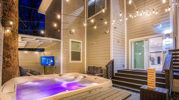 A hot tub on a wooden deck with string lights, a staircase leading to a door, and a TV screen on the wall. - Stylish 6BR/5BA, DT, Hot Tub, Games, Sleeps 20+ (San Antonio)