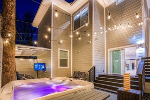 Een hottub op een houten terras, versierd met lichtjes, met een trap naar een deur en een televisiescherm aan de muur.