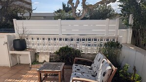Eine Terrasse mit weißem Gitterzaun, einem Holztisch, einer gepolsterten Bank und Kübelpflanzen.