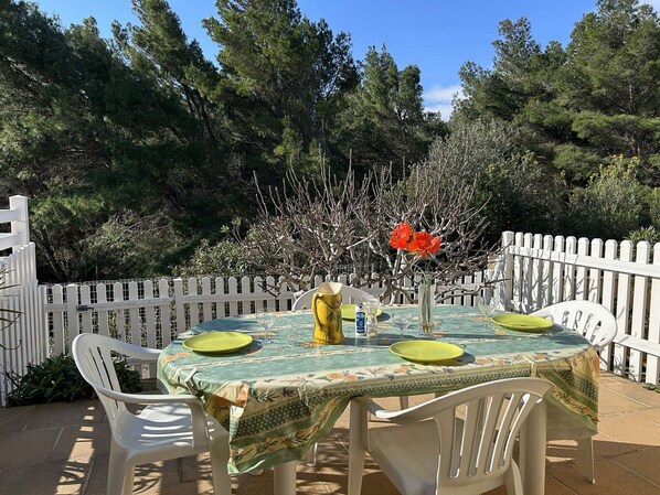 Un área de comedor al aire libre con una mesa dispuesta para cuatro personas, rodeada de sillas blancas, una cerca de piquetes blancos y árboles densos al fondo.