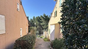 Un camí de grava estret flanquejat per edificis amb persianes i un costat arbrat amb setge.
