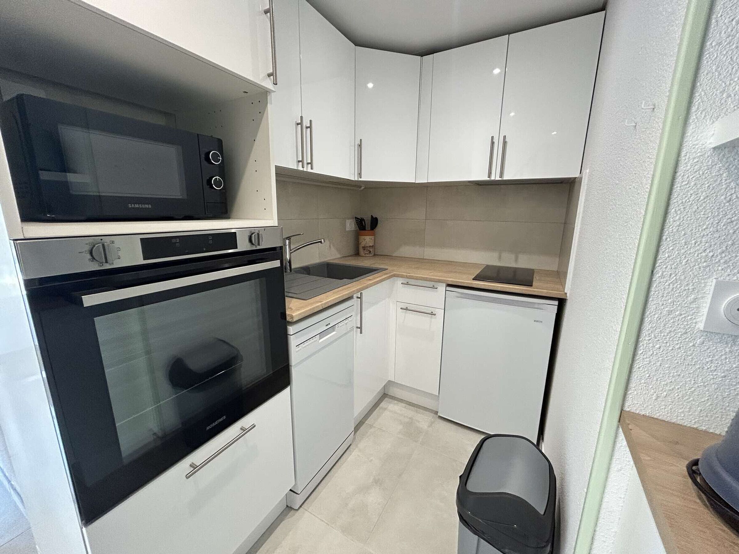 Een kleine keuken met witte kastjes, een magnetron, oven, vaatwasser en een spoelbak.
