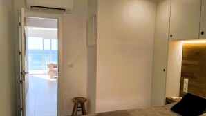 Ein Schlafzimmer mit einem Bett, einem hölzernen Hocker, einem Nachttisch und Blick aufs Meer durch eine offene Tür.