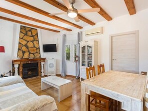 A living room with a stone fireplace, a television, a dining table with chairs, and a ceiling fan. - Villa with private pool, near beach - 6 guests (Oliva)