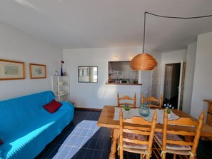 Un salon avec un canapé bleu, une table à manger en bois et des chaises. Il y a également un coin cuisine avec un luminaire suspendu.