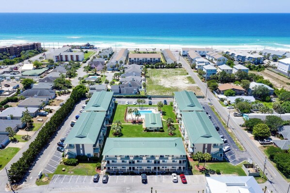 Vue aérienne d’un hôtel situé sur la côte, avec une piscine centrale, entouré de bâtiments d’un seul étage et une plage en arrière-plan.